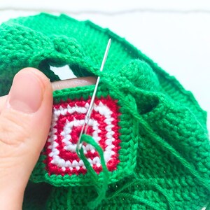 May include: Close-up of a green crocheted hat with a red and white crocheted square design. A needle and thread are being used to sew the square onto the hat.
