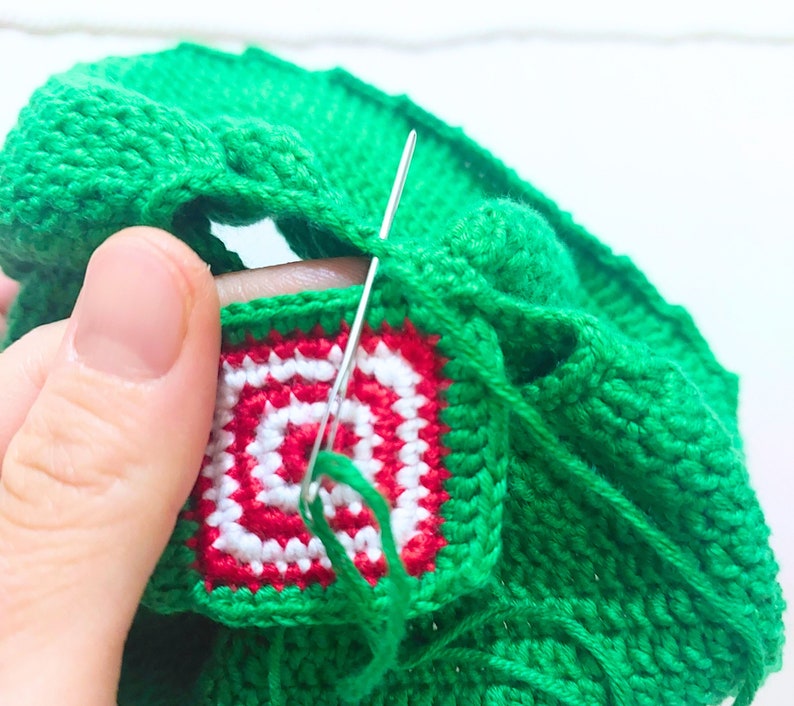 May include: Close-up of a green crocheted hat with a red and white crocheted square design. A needle and thread are being used to sew the square onto the hat.