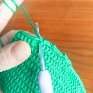May include: Close-up of a person crocheting a green knitted item with a white crochet hook. The yarn is green and the hook is being used to create a stitch.