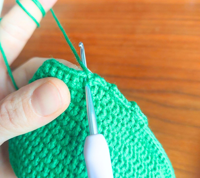 May include: Close-up of a person crocheting a green knitted item with a white crochet hook. The yarn is green and the hook is being used to create a stitch.