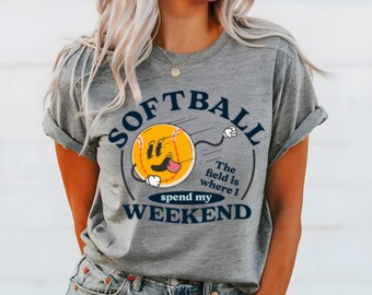 May include: Gray t-shirt with a graphic of a cartoon softball with a tongue sticking out. The text "Softball" is in a blue arc above the softball. The text "The field is where I spend my weekend" is below the softball.