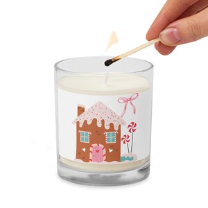 Könnte beinhalten: Eine weiße Kerze in einem durchsichtigen Glasgefäß mit einem Lebkuchenhaus-Design. Das Haus ist mit rosa Zuckerguss, Streuseln und Zuckerstangen verziert. Die Kerze wird mit einem Streichholz angezündet.