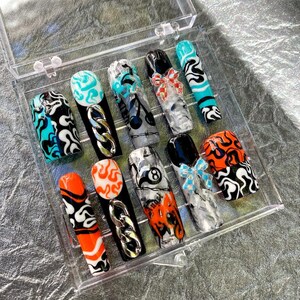 May include: A clear plastic box containing ten press-on nails with various designs. The nails feature black and white flames, a chain link, a black and white 8-ball, a black and white cherry, a black and white marble design, and a blue bow. The nails are in a variety of colors, including orange, black, white, and blue.