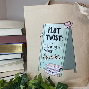 May include: A beige canvas tote bag featuring a green and white sign that reads "Plot Twist: I bought more books." The sign has a pink flower on top and a stack of books underneath.