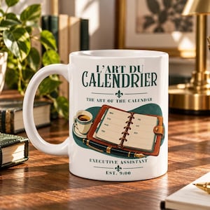 Op de afbeelding: Een witte keramische mok met de tekst L'ART DU CALENDRIER, THE ART OF THE CALENDAR, EXECUTIVE ASSISTANT en EST. 9:00. De afbeelding toont een open bruine lederen agenda, een vulpen en een klein kopje koffie op een blauwgroene achtergrond.