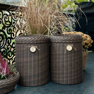 Könnte beinhalten: Zwei braune Pflanzenkörbe aus Weide mit geflochtenem Muster. Die Körbe stehen auf einer Terrasse mit einem dekorativen Metallgeländer im Hintergrund. Die Körbe haben kleine Holzschildchen mit dem Text "Garden Plants" und "Plants & Flowers" darauf.