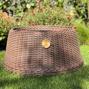 May include: A round, brown, woven wicker tree collar with a tapered shape and a wide opening. A small, circular, light-colored wooden tag is attached to the front, featuring the text Garden & Home Decor printed in a dark brown font.