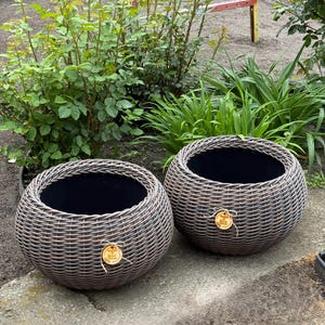 Può includere: Due fioriere rotonde marroni intrecciate con interno scuro sono su una superficie di cemento. Le fioriere hanno un aspetto naturale e strutturato, e ognuna ha una piccola etichetta di legno attaccata con spago. Piante verdi e fogliame sullo sfondo.