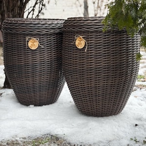 Può includere: Due cesti portaoggetti in vimini intrecciato marrone scuro con coperchi. Ogni cesto ha un'etichetta rotonda in legno attaccata con spago. I cesti sono all'aperto, sulla neve e sull'erba, con alberi sullo sfondo.