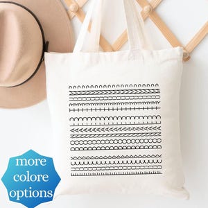 May include: A beige canvas tote bag featuring a black graphic print of various horizontal line patterns and a tan felt hat hanging on a wooden accordion rack. A blue star-shaped badge with the text more colore options is visible.