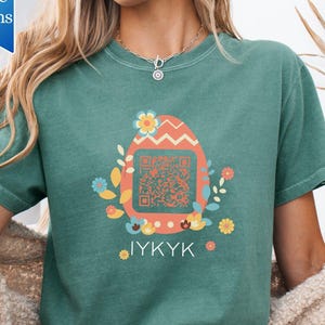 May include: Teal green t-shirt featuring a colorful Easter egg design with a QR code and floral accents. The shirt has the text "IYKYK" below the egg. A small blue banner in the upper left corner says "more color options".