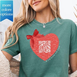 May include: Teal-colored t-shirt featuring a red heart design with a QR code and a red bow. The heart is filled with the word "love" and the phrase "If you know you know". The shirt has a relaxed fit.