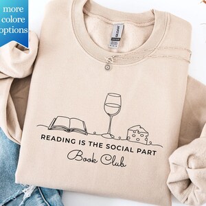 May include: Beige crewneck sweatshirt with a black line drawing of a book, wine glass, and cheese. The text "READING IS THE SOCIAL PART" and "Book Club" are below the graphic. A silver necklace with a sunflower pendant is on the sweatshirt.