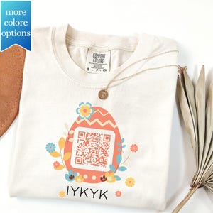 May include: Cream-colored t-shirt featuring a coral Easter egg design with a QR code, surrounded by floral accents. The text "IYKYK" is printed below the egg. A gold necklace and a decorative leaf are also present.