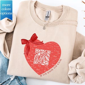 May include: A beige sweatshirt featuring a large red heart design with a QR code and the words "love" and "If you know you know." A red bow is on top of the heart. A silver necklace is visible.