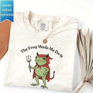 May include: Cream-colored t-shirt featuring a cartoon frog dressed as a devil, holding a pitchfork. The text "The Frog Made Me Do It" is printed above the frog. A gold necklace with a pendant is draped on the shirt.
