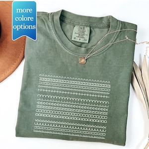 May include: An olive green t-shirt with a white graphic of various stitch patterns. The shirt has a crew neck and short sleeves. A gold necklace with a pendant rests on the shirt. The tag reads "COMFORT COLORS". The text "more colore options" is in a blue box.
