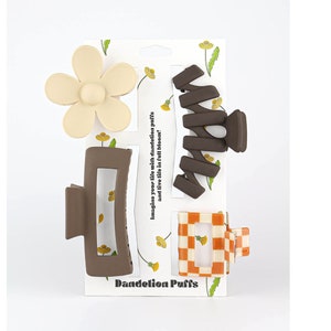 May include: A selection of hair clips in different styles, including a flower, a rectangular clip, a spiral clip, and a checkered clip. The clips are in shades of brown, beige, and orange. The packaging reads "Dandelion Puffs" and "Imagine your life with dandelion puffs and live life in full bloom!"