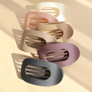 Könnte beinhalten: Eine Ansammlung von sechs ovalen Haarspangen mit matter Oberfläche, die in einem Stapel angeordnet sind. Die Spangen verfügen über ein ausgeschnittenes Design mit horizontalen Streben in den Farben Creme, Altrosa, Beige, Sand, Mauve und Anthrazit.