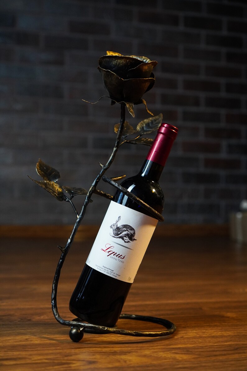 May include: A black metal wine bottle holder shaped like a rose with a bottle of red wine inside. The label on the bottle says "Lquus Vinho Tinto".