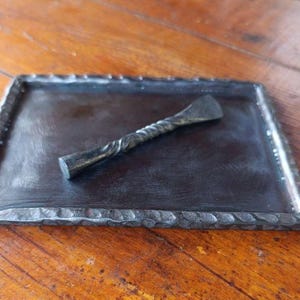 May include: A rectangular dark gray metal tray with a textured, hammered edge and a matching metal spreader tool featuring a twisted handle design. The tray has a smooth, flat center surface, while the spreader has a flat, angled blade end.