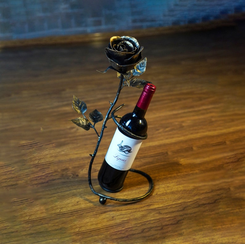 May include: A black metal wine bottle holder shaped like a rose with a bottle of red wine inside. The rose has gold accents and is on a wooden surface.