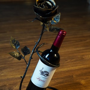 May include: A black metal wine bottle holder shaped like a rose with a bottle of red wine inside. The label on the bottle reads "Lupus Vino Tinto".