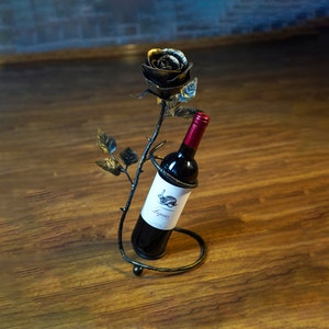 May include: A black metal wine bottle holder shaped like a rose with a bottle of red wine inside. The rose has gold accents and is on a wooden surface.