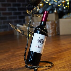 May include: A black metal wine bottle holder shaped like a rose with a bottle of red wine inside. The label on the bottle reads "Lepus Vino Tinto".