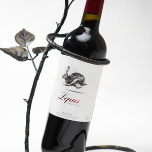May include: A black metal wine bottle holder shaped like a rose bush with a bottle of red wine inside. The bottle label reads "Lepus Vinho Tinto".