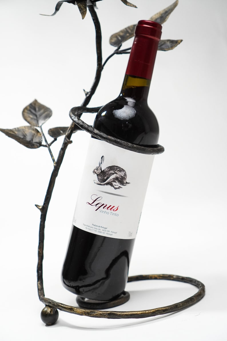 May include: A black metal wine bottle holder shaped like a rose bush with a bottle of red wine inside. The bottle label reads "Lepus Vinho Tinto".