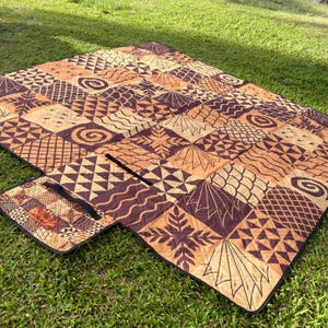 May include: A quilted outdoor blanket featuring a patchwork design of brown and tan squares with various geometric, spiral, and leaf-like patterns. The blanket includes a foldable flap with a black handle for carrying and a small rectangular label.