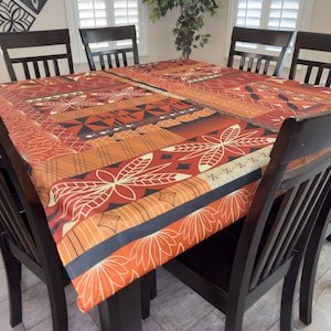 May include: A rectangular dining table with a colourful patterned tablecloth featuring geometric shapes and floral designs in shades of orange, red, and black. The table is surrounded by four black chairs.