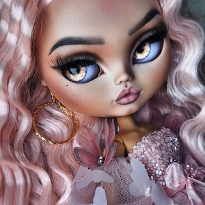 May include: A close-up of a fashion doll with pink wavy hair and large, expressive eyes. The doll has full lips, long eyelashes, and gold hoop earrings. She wears a pink dress adorned with sequins and butterfly accents, creating a glamorous aesthetic.