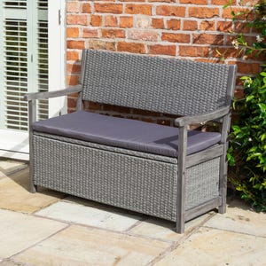 May include: A grey wicker storage bench with a grey cushion and wooden arms. The bench is sitting on a patio.