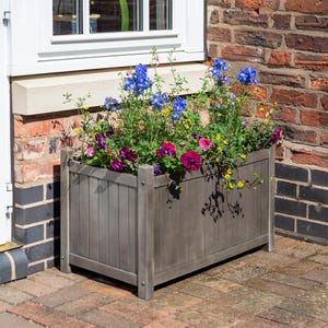 May include: A grey wooden planter box filled with colourful flowers, including blue, pink, and yellow blooms. The planter is placed on a brick patio next to a brick wall and a white window.