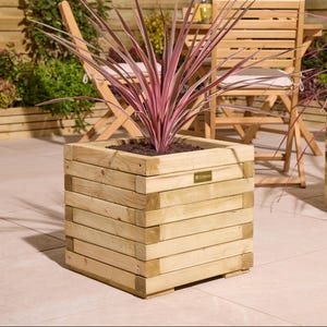 May include: A square wooden planter with a layered design, containing soil and a plant with long, pink and green leaves. The planter is made of light-coloured wood and has a small metal plate on the front.