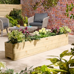 May include: A long, rectangular wooden planter filled with a variety of flowers. The light-coloured wood has a natural finish. The flowers display green, yellow, and purple hues. The planter sits on a patio, with garden furniture visible in the background.