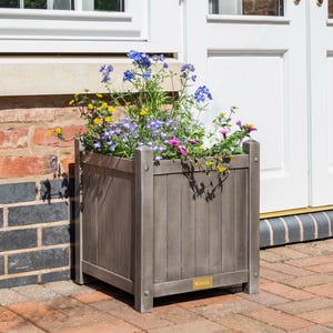 May include: A grey wooden planter box filled with a variety of colourful flowers, including blue, purple, pink and yellow. The planter is sitting on a brick patio.