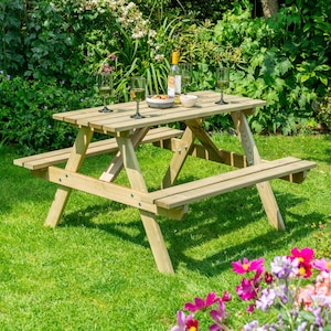 May include: A light brown wooden picnic table with attached benches, set on a green lawn. The table has a rectangular top. Wine glasses, a bottle of wine, and snacks are on the table. The background features green foliage and colourful flowers.