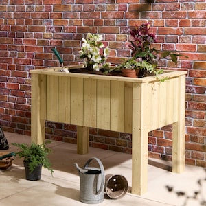 May include: A rectangular raised garden bed made of light-coloured natural wood with four sturdy legs. The planter contains soil, white hellebore flowers, and a dark pink flowering plant. A metal watering can sits on the ground nearby.
