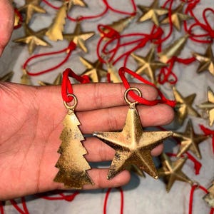 Puede incluir: Una colección de adornos de metal en tono dorado con formas de estrellas de cinco puntas y árboles de Navidad. Cada adorno presenta un acabado desgastado y envejecido, y está unido a un cordón rojo brillante para colgarlo fácilmente.