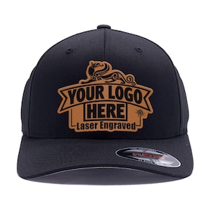 May include: A black baseball cap featuring a tan leather-like patch on the front. The patch displays a winged creature graphic above a banner with the text YOUR LOGO HERE and the words Laser Engraved below. A circular sticker is on the brim.