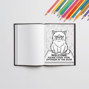 May include: An open coloring book with a black cover, featuring a black and white illustration of a grumpy cat. The text below the cat reads "WELCOME. PLEASE LEAVE YOUR OPTIMISM AT THE DOOR." Colored pencils are scattered around the book.