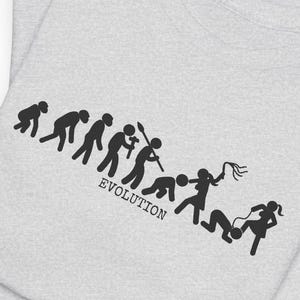 May include: A light gray t-shirt featuring a black graphic print that depicts a sequence of figures in an evolution progression, ending with a figure holding a whip over another figure, with the word EVOLUTION printed below the figures.