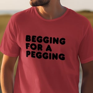 May include: Red t-shirt with the phrase "BEGGING FOR A PEGGING" in black, bold letters. The shirt is short-sleeved and appears to be made of a soft material. The background shows a beach and grassy area.