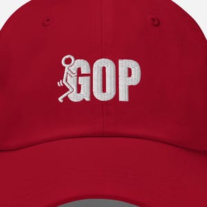 Könnte beinhalten: Rote Baseballkappe mit dem weiß gestickten Wort "GOP". Das "O" in "GOP" wird durch eine Strichmännchen-Figur ersetzt. Die Kappe hat einen gebogenen Schirm und einen verstellbaren Riemen.