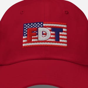 May include: A red baseball cap featuring an embroidered rectangular patch on the front. The patch displays a stylised American flag design with the letters FDT stitched in bold, block-style font across the centre in red, white, and blue.