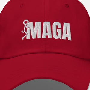 Könnte beinhalten: Rote Baseballkappe mit weiß gesticktem Text und Grafik. Der Text lautet "MAGA" mit einer stilisierten Figur. Die Kappe hat einen gebogenen Schirm und einen Stoffriemenverschluss.