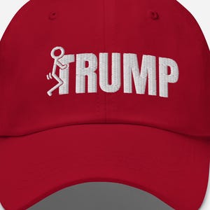 Könnte beinhalten: Rote Baseballkappe mit weiß gesticktem Schriftzug "TRUMP". Der Buchstabe "F" ist durch eine Strichmännchen-Figur ersetzt. Die Kappe hat einen gebogenen Schirm und einen verstellbaren Verschluss.